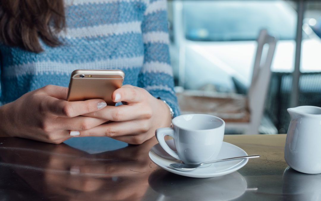 Close-up of woman hands and torso as she sits at table in a public restaurant. The table is set with coffee cup and small creamer pitch. She is text messaging with her cell phone.