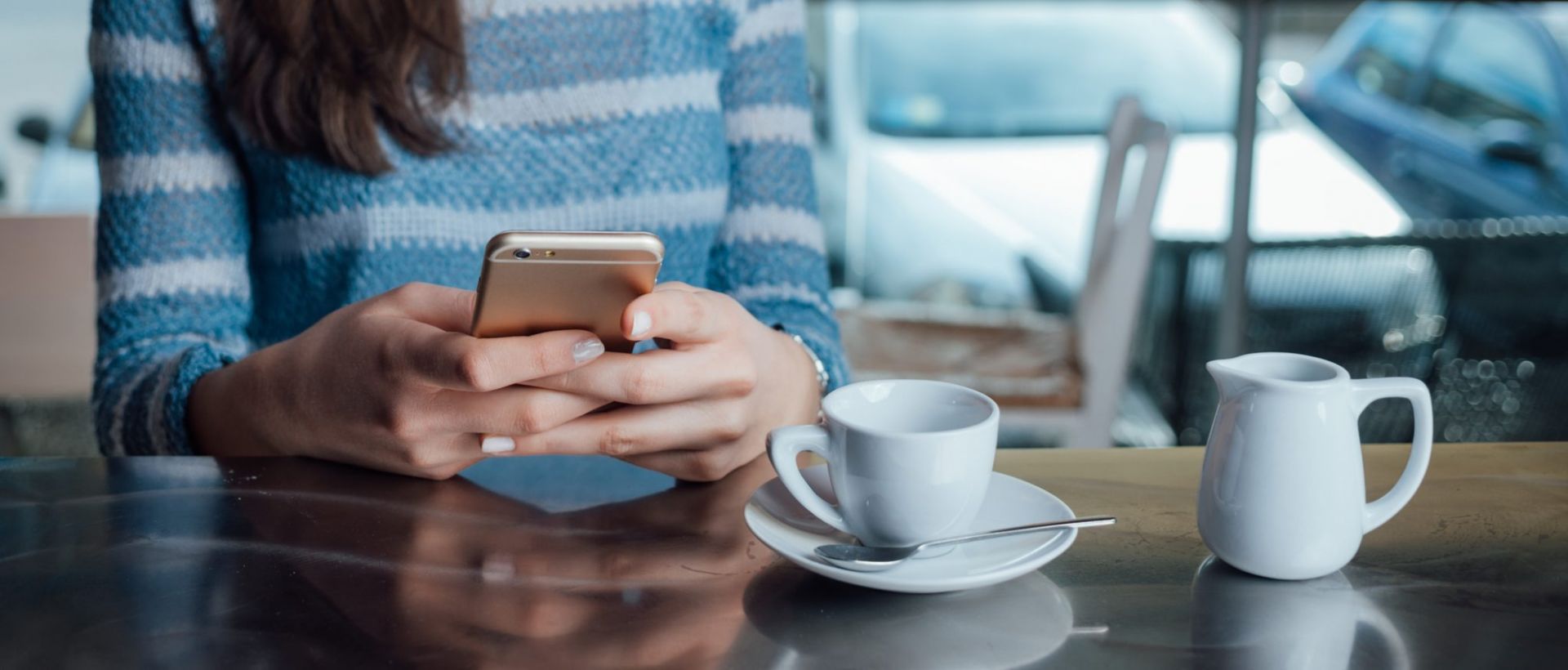 Close-up of woman hands and torso as she sits at table in a public restaurant. The table is set with coffee cup and small creamer pitch. She is text messaging with her cell phone.